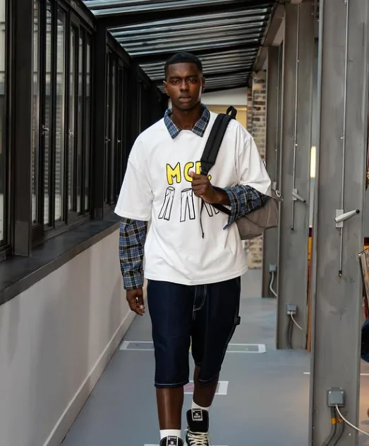 Un jeune homme en t-shirt blanc MCD, chemise à carreaux et bermuda en jean marche dans une coursive vitrée.